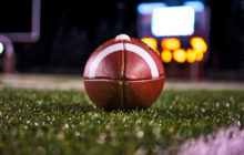 This is an image of a football on a field at night with the scoreboard and goal posts in the background.